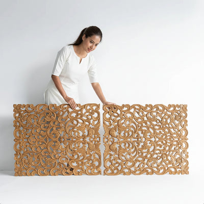A woman in a white t-shirt and white pants holds up two large, ornate, hand-carved wooden screens with intricate scrollwork patterns, set against a plain white studio background.
